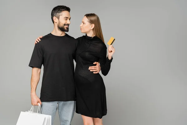 Hombre barbudo sonriente sosteniendo bolsas de compras y abrazando a mujer embarazada con estilo en vestido negro sosteniendo tarjeta de crédito aislada en gris, nuevos comienzos y el concepto de crianza, marido y mujer - foto de stock