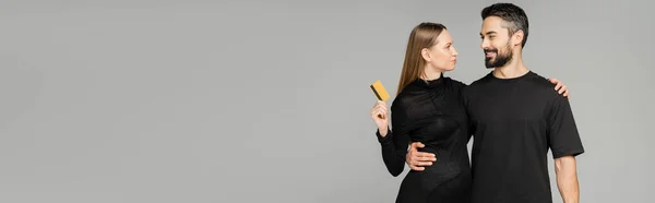 Lächelnder bärtiger Mann im schwarzen T-Shirt umarmt schwangere Frau in stylischem Kleid und hält Kreditkarte in der Hand, während er isoliert auf grauem, Neuanfang und Erziehungskonzept steht, Banner — Stockfoto