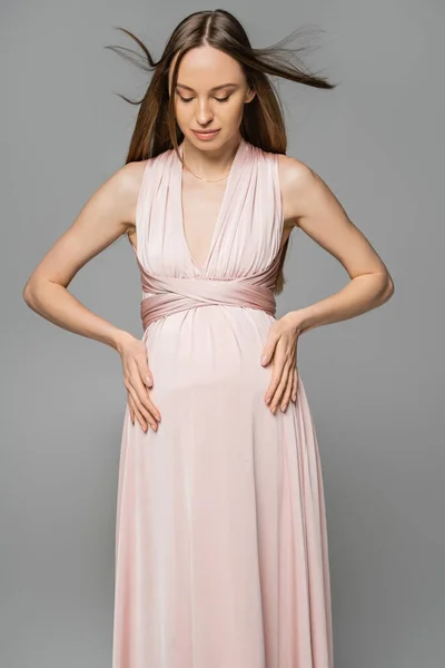 Модная и длинноволосая беременная женщина в розовом платье касается живота и смотрит вниз, стоя изолированным на серой, элегантной и стильной одежде беременности, чувственности, будущей матери — стоковое фото