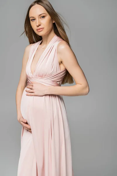 Модная и красивая беременная женщина в розовом платье, смотрящая в камеру, стоя и позируя в изоляции на сером, элегантном и стильном наряде для беременности, чувственности, будущей матери — стоковое фото