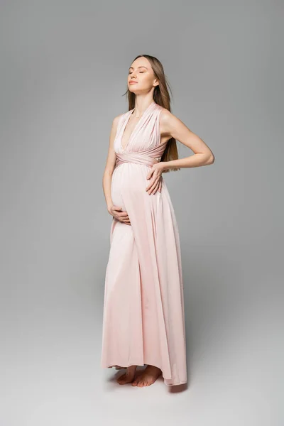 Полная длина расслабленной и светловолосой беременной женщины в розовом платье касаясь живота, стоя на сером фоне, элегантный и стильный наряд беременности, чувственность, будущая мать — стоковое фото