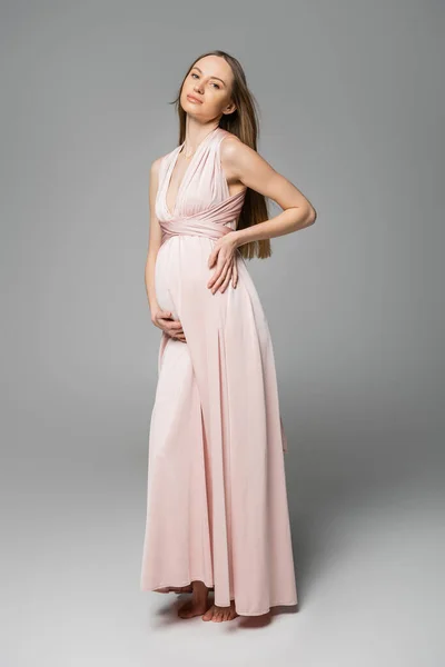 Mulher grávida de cabelos finos e na moda em vestido rosa olhando para a câmera enquanto posando em fundo cinza, traje de gravidez elegante e elegante, sensualidade, futura mãe — Fotografia de Stock