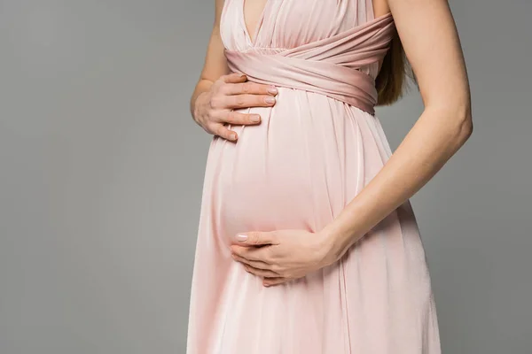 Обрезанный вид на модную и длинноволосатую женщину в розовом платье, касающуюся живота, стоящую изолированную на серой, элегантной и стильной одежде для беременности, чувственности, будущей матери — стоковое фото