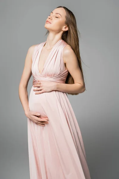 Розслаблена прекрасна волохата і очікувана мати в рожевій сукні торкається живота, одночасно позує і стоїть ізольовано на сірому, елегантному і стильному одязі вагітності, чуттєвості, матері-до-бути — стокове фото
