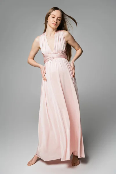 Повна довжина босоніж довгошерстої і вагітної жінки в рожевому платті, що торкається живота, стоячи на сірому фоні, елегантний і стильний одяг для вагітності, чуттєвість, мати-до-бути — стокове фото