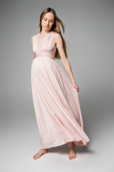 Madre embarazada descalza y de moda en vestido rosa posando y tocando tela sobre fondo gris, elegante y elegante atuendo de embarazo, sensualidad, futura madre - foto de stock