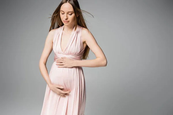 Mulher grávida de cabelos finos na moda posando em vestido rosa e barriga tocante enquanto está isolada em cinza com espaço de cópia, traje de gravidez elegante e elegante, sensualidade, futura mãe — Fotografia de Stock