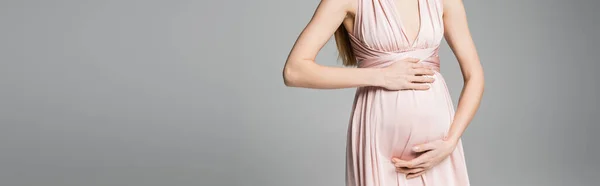 Vue recadrée de la femme à la mode et enceinte en robe rose touchant le ventre tout en se tenant isolé sur gris avec espace de copie, tenue de grossesse élégante et élégante, bannière — Photo de stock