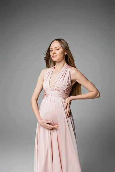 Femme à la mode aux cheveux longs et enceinte avec les yeux fermés touchant le ventre tout en posant en robe rose isolé sur gris, tenue de grossesse élégante et élégante, sensualité, future mère — Photo de stock