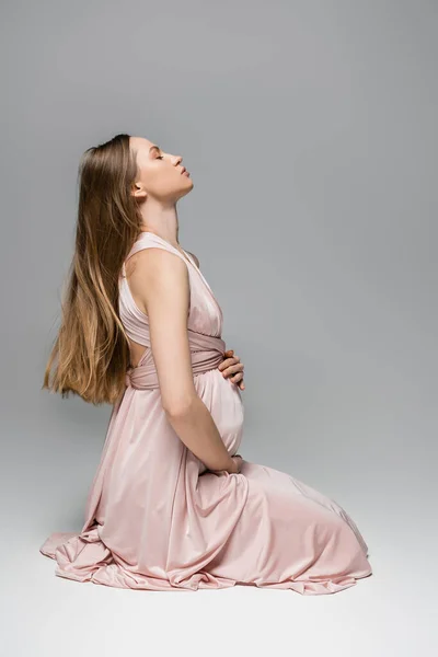 Vue latérale des cheveux longs attendant mère en robe rose tendance touchant le ventre tout en étant assis sur fond gris, tenue de grossesse élégante et élégante, sensualité, future mère — Photo de stock
