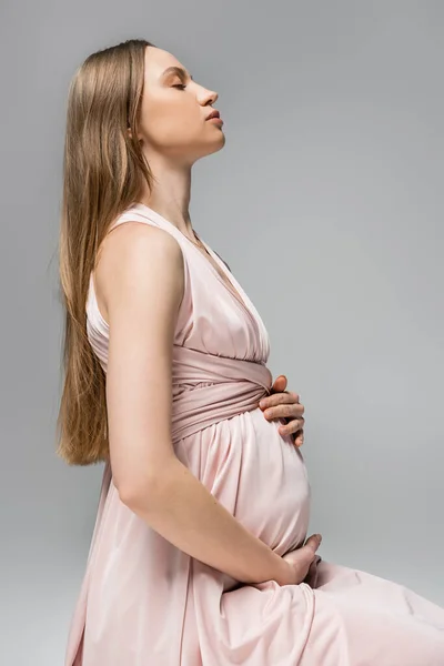 Вид сбоку расслабленной и беременной женщины с натуральным макияжем, касающимся живота, позируя в розовом платье и сидя изолированно на серой, элегантной и стильной одежде для беременности, чувственности, будущей матери — стоковое фото