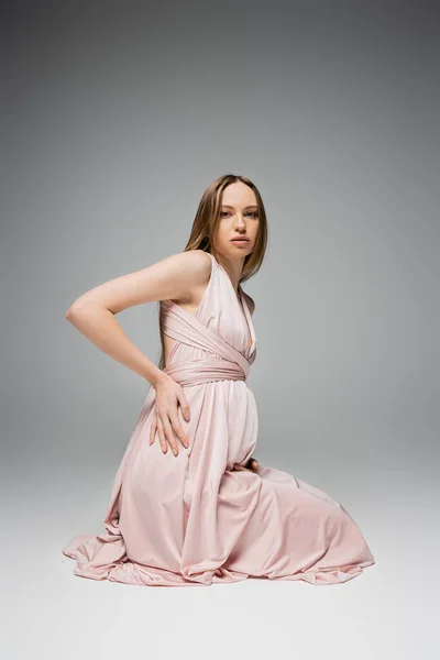 Модна і справедлива вагітна жінка з натуральним макіяжем в рожевій сукні, дивлячись на камеру, сидячи на сірому фоні, елегантний і стильний одяг для вагітності, чуттєвість, мати-бути — стокове фото