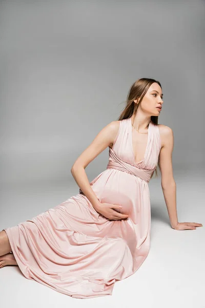Модная будущая мама в розовом платье трогает животик, расслабляясь, отводя взгляд и сидя на сером фоне, чувственность, будущая мама, элегантный и стильный наряд беременности — стоковое фото
