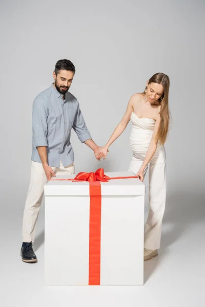 Full length of stylish couple holding hands while standing together near bog gift box during gender reveal surprise party and celebration on grey background, expecting parents concept — Stock Photo