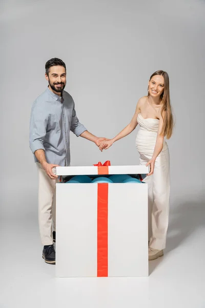 Весела і стильна пара тримає руки, відкриваючи болото подарункової коробки з синіми кульками під час статі розкриває сюрприз вечірку на сірому фоні, очікуючи від батьків концепції, це хлопчик — стокове фото