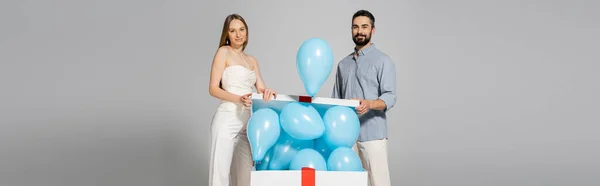 Smiling and trendy expecting parents looking at camera while opening bog gift box with festive blue balloons during gender reveal surprise party isolated on grey, it`s a boy, banner — Stock Photo