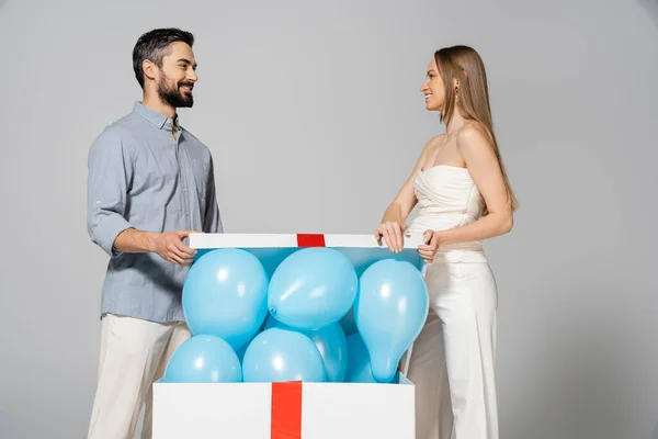 Cheerful and stylish pregnant woman looking at husband while opening big gift box with festive blue balloons during gender reveal surprise party isolated on grey, expecting parents, it`s a boy — Stock Photo