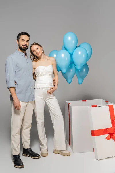 Voller positiver werdender Eltern, die sich umarmen und in die Kamera schauen, in der Nähe einer großen Geschenkbox mit festlich blauen Luftballons während der Geschlechterparty auf grauem Hintergrund, es ist ein Junge — Stockfoto