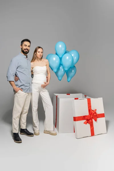 Full length of trendy and cheerful expecting parents hugging each other while standing near blue balloons and big gift box during gender reveal surprise party on grey background, it`s a boy — Stock Photo