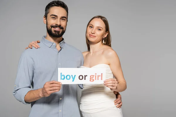 Positive and trendy pregnant woman holding card with boy or girl lettering and hugging stylish husband during gender reveal surprise party isolated on grey, expecting parents concept — Stock Photo