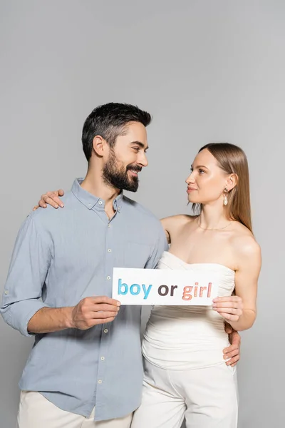 Positive and bearded man holding card with boy or girl lettering and hugging stylish pregnant wife during celebration and gender reveal surprise party isolated on grey, expecting parents concept — Stock Photo
