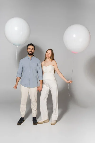 Full length of positive and stylish expecting parents looking at camera while holding hands and balloons during gender reveal surprise party on grey background, expecting parents concept — Stock Photo