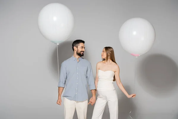 Positive and trendy expecting parents holding hands and festive balloons during celebration and gender reveal surprise party on grey background, expecting parents concept — Stock Photo