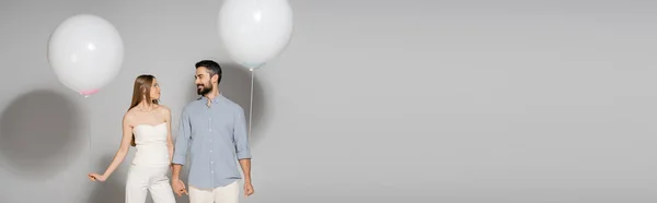 Модная беременная женщина, держащая за руку улыбающегося мужа и праздничный воздушный шар во время гендерного раскрытия сюрприз-вечеринки на сером фоне с копировальным пространством, баннером, ожидающей от родителей концепции — стоковое фото