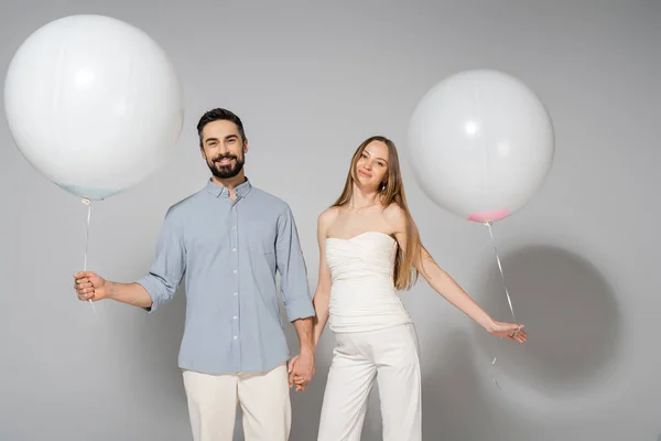 Lächelnde und stylische werdende Eltern, die Händchen halten und festlich weiße Luftballons, während sie beim Geschlechtsverkehr in die Kamera schauen, verraten Überraschungsparty auf grauem Hintergrund — Stockfoto