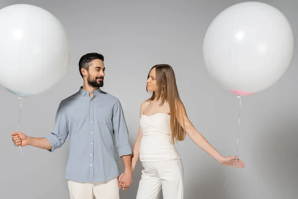 Усміхнені і модні очікувані батьки тримають руки і білі святкові кульки, дивлячись один на одного під час святкування і гендер розкривають сюрприз вечірку на сірому фоні — стокове фото