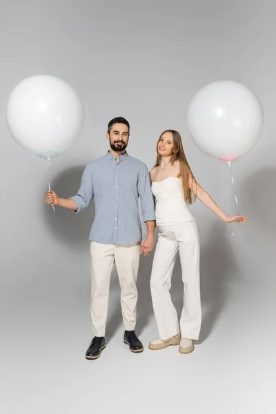 Full length of fashionable and happy expecting parents holding white balloons and looking at camera during gender reveal surprise party on grey background — Stock Photo
