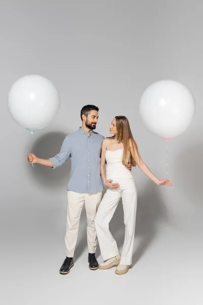 Full length of fashionable expecting parents hugging and looking at each other while holding festive balloons during gender reveal surprise party on grey background — Stock Photo