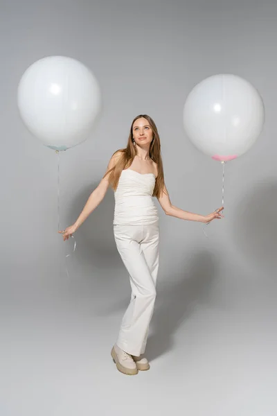 Full length of trendy and pregnant woman looking away while holding white festive balloons during celebration and gender reveal surprise party on grey background, fashionable pregnancy attire — Stock Photo