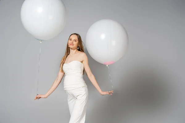 Позитивна і модна вагітна жінка, дивлячись на камеру, тримає білі святкові кульки під час статі, розкриває сюрприз вечірку на сірому фоні, модний одяг для вагітності — стокове фото