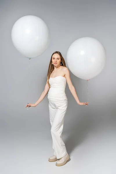 Повна довжина модної і красивої вагітної жінки, що тримає білі кульки і дивиться на камеру під час статі, розкриває сюрприз вечірку на сірому фоні, модний одяг для вагітності — стокове фото