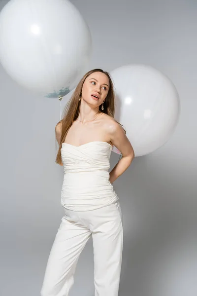 Modische schwangere Frau, die wegschaut, während sie festliche weiße Luftballons in der Hand hält und beim Geschlechtsverkehr steht, verrät Überraschungsparty auf grauem Hintergrund, modische Schwangerschaftskleidung — Stockfoto