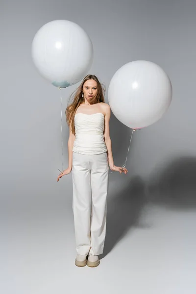 Full length of stylish and shocked expecting mother lookin at camera while holding white festive balloons during gender reveal surprise party on grey background, fashionable pregnancy attire — Stock Photo