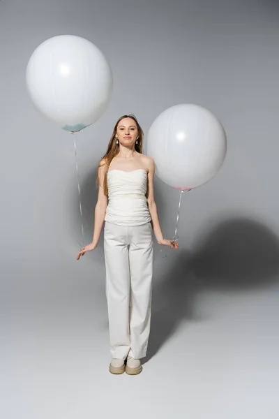 Full length of pleased and fashionable pregnant woman looking at camera while holding white balloons during gender reveal surprise party on grey background, fashionable pregnancy attire — Stock Photo