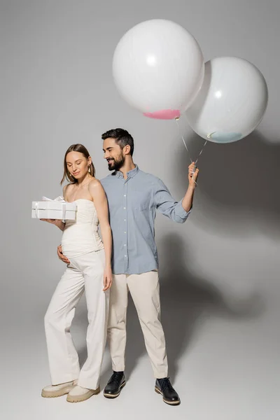 Веселий чоловік обіймає стильну вагітну дружину і тримає подарункову коробку і святкові кульки під час святкування і статі розкриває сюрприз вечірку на сірому фоні, модний одяг для вагітності, хлопчик або дівчинка — стокове фото