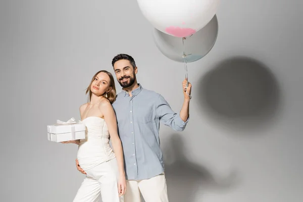 Positive and stylish man holding festive balloons and hugging pregnant wife with gift box during celebration and gender reveal surprise party on grey background, pregnancy attire, boy or girl — Stock Photo
