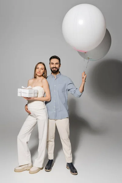 Повна довжина позитивного чоловіка, який тримає святкові кульки і обіймає модну і вагітну дружину з подарунковою коробкою, стоячи разом під час дитячої вечірки на сірому фоні, хлопчика або дівчинки — стокове фото