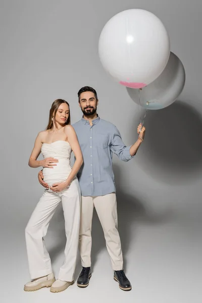 Volle Länge der positive und stilvolle Mann hält festliche Luftballons und umarmt schwangere Frau während Baby-Dusche Party auf grauem Hintergrund, erwartete Eltern Konzept, Junge oder Mädchen — Stockfoto