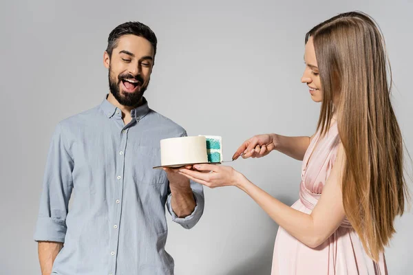 Задоволений і бородатий чоловік тримає торт біля веселої вагітної дружини з ножем під час дитячої вечірки для душу на сірому фоні, очікуючи від батьків концепцію, гендерну вечірку, це хлопчик — стокове фото