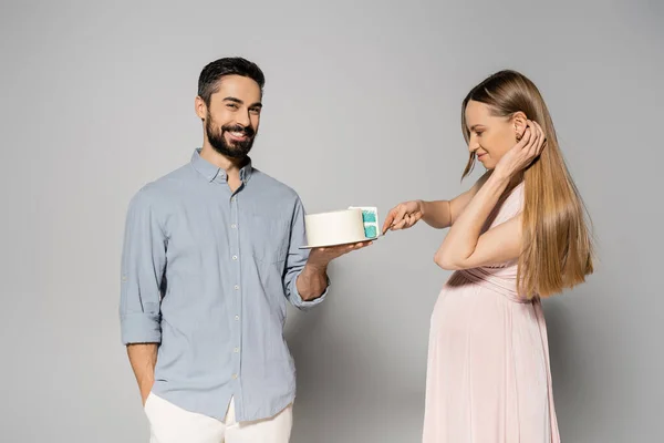 Positive and bearded man holding cake near elegant pregnant wife in pink dress during gender party on grey background, expecting parents concept, gender party, it`s a boy, cutting cake — Stock Photo