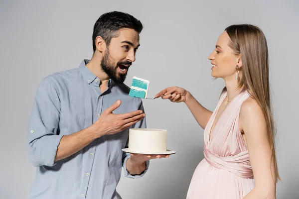 Улыбающаяся беременная женщина в розовом платье держит синий торт рядом с мужем с открытым ртом во время празднования гендерной вечеринки на сером фоне, ожидая концепцию родителей, это мальчик — стоковое фото