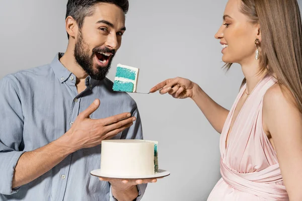 Усміхнена і елегантна вагітна жінка годує чоловіка синім тортом під час святкування гендерної вечірки ізольовано на сірому, очікуючи, що батьки концепції щастя, це хлопчик — стокове фото