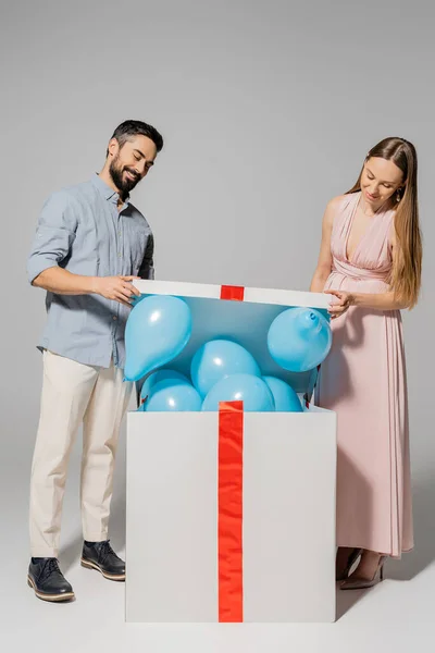 Positiva y elegante pareja abriendo una gran caja de regalo con globos azules durante la ducha del bebé sobre fondo gris, esperando el concepto de los padres, fiesta de género, es un niño - foto de stock