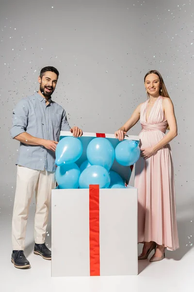 Longitud completa de la pareja de moda mirando a la cámara al abrir la caja de regalo con globos azules y de pie debajo de confeti durante la ducha del bebé sobre fondo gris, fiesta de género, es un niño - foto de stock
