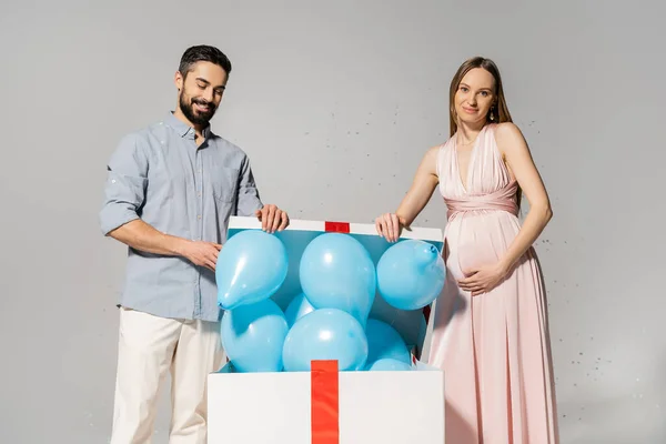 Caja de regalo de apertura de pareja positiva y elegante con globos azules cerca de confeti festivo mientras celebra la fiesta de la ducha del bebé sobre fondo gris, esperando el concepto de los padres, fiesta de género, es un niño - foto de stock