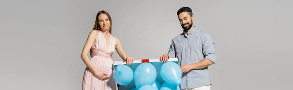 Елегантна і вагітна жінка відкриває подарункову коробку з блакитними повітряними кулями біля веселого чоловіка і конфетті під час святкування дитячого душу на сірому фоні, гендерної вечірки, це хлопчик, банер — стокове фото